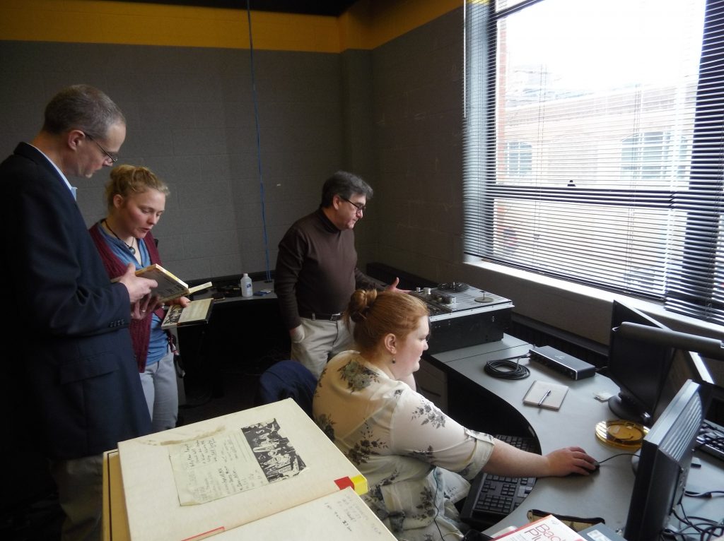 Four people working. Two reading a book, one on a computer, and another at a reel to reel machine.