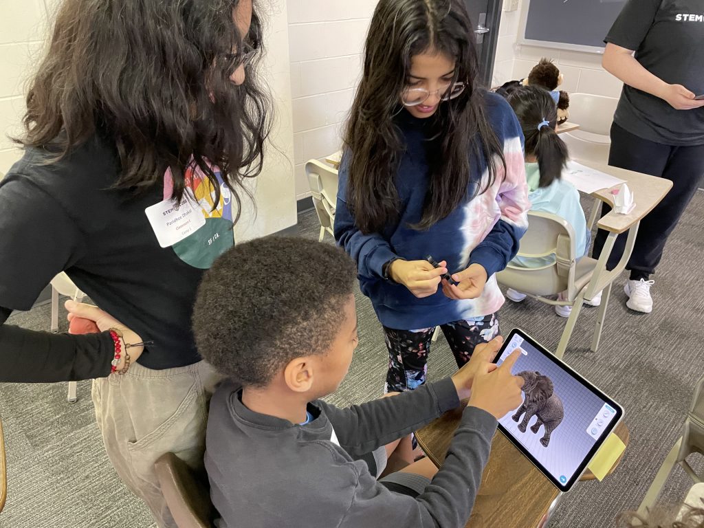 Younger kids and Mizzou students study a 3D model on a tablet.